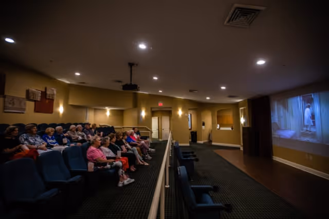 A group of elderly people seated in a small theater room watching a movie projected on a screen. The room has dim lighting, carpeted floors, and rows of comfortable theater seats.