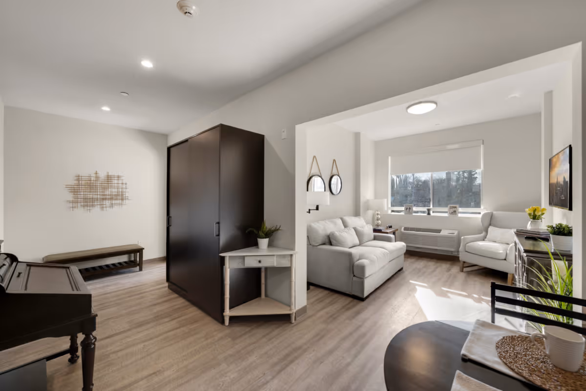 Bright furnished living area with a sofa and armchair by a large window, a small dining table, and a dark wardrobe dividing the space.