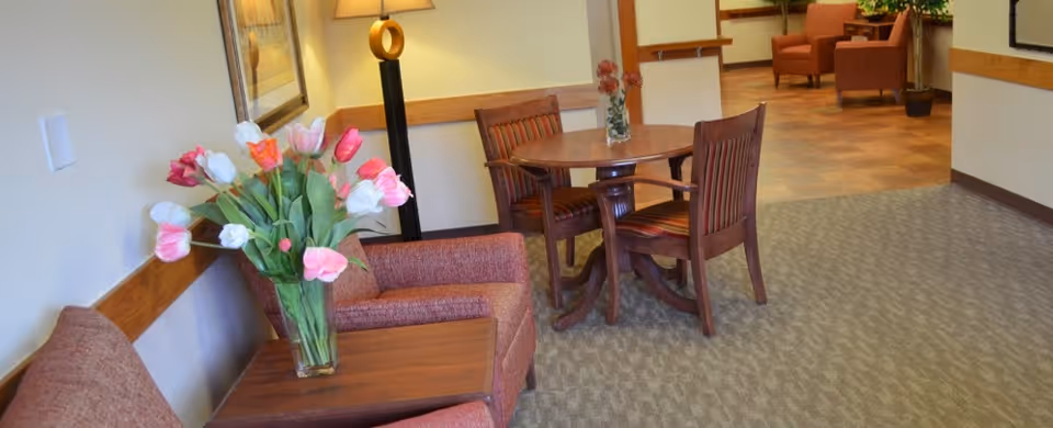 A cozy seating area in a senior living facility with two upholstered armchairs, a wooden side table with a vase of colorful tulips, a round wooden table with two wooden chairs, a floor lamp, and additional seating visible in the background.