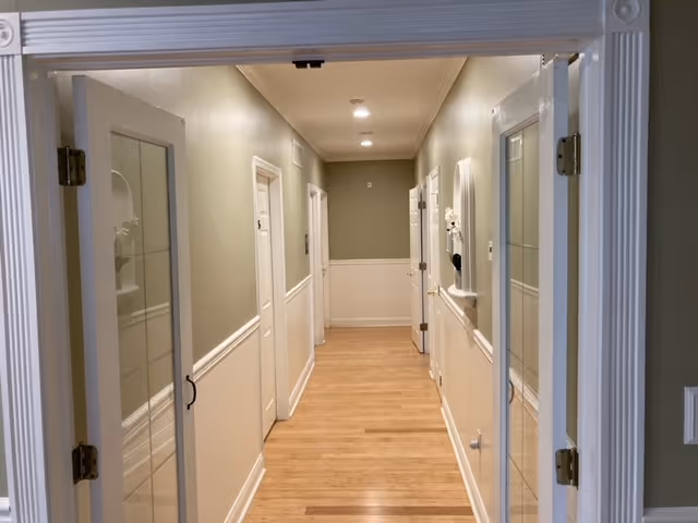 View down a clean, well-lit hallway with light wood flooring and light green walls with white wainscoting. Several white doors line the hallway on both sides, and there are recessed ceiling lights. The hallway is framed by a white door frame with glass panels on either side.
