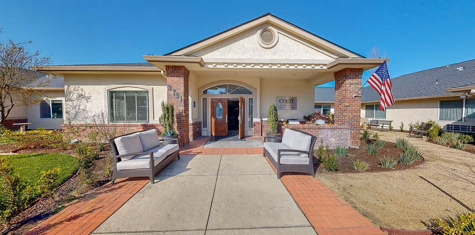 Front entrance of Cogir of Turlock senior living facility with two cushioned outdoor sofas on either side of the walkway, brick pillars, an American flag, and landscaped garden areas under a clear blue sky.