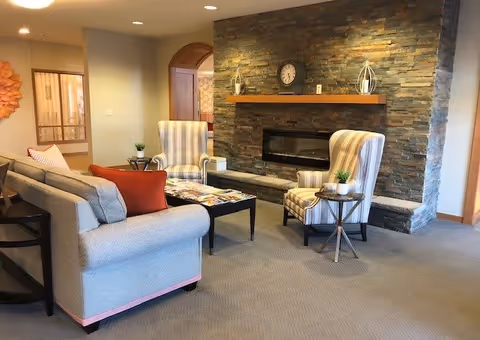 A cozy living room area with a gray sofa adorned with orange and gray pillows, two striped armchairs, a stone fireplace with a wooden mantle holding decorative items and a clock, and a small round side table with a plant. The room has beige carpet and soft lighting.