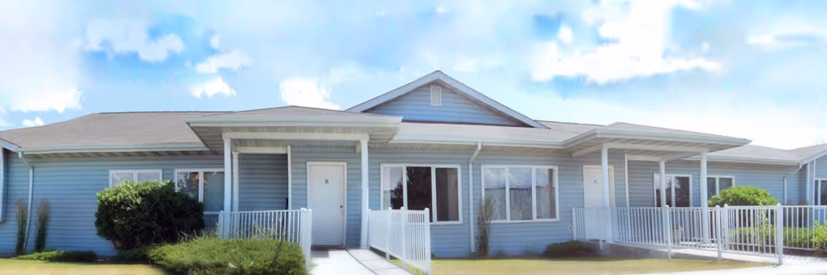 Exterior front view of a single-story building with light blue siding and white trim, featuring multiple entrances with small covered porches and white railings, surrounded by green bushes and grass under a partly cloudy sky.