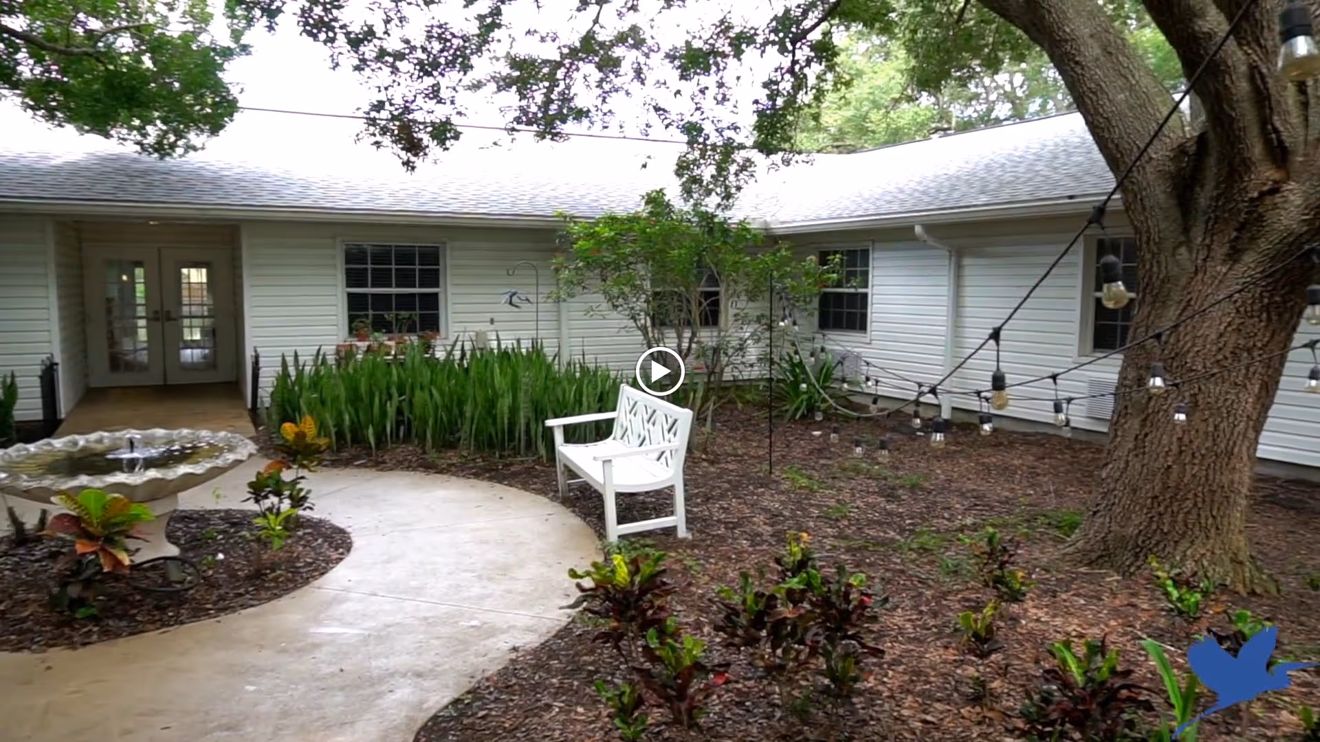 Outdoor courtyard area of a senior living facility with a curved concrete pathway, a white bench, a birdbath fountain, various plants, and string lights hanging from a large tree. The building with white siding and multiple windows surrounds the courtyard.