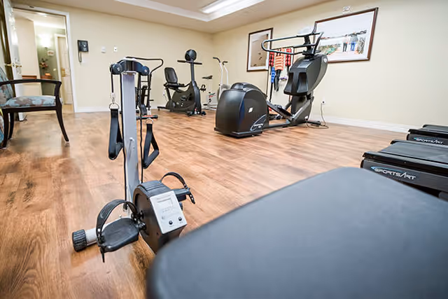 Interior view of a fitness room with various exercise equipment including stationary bikes, an elliptical machine, and a rowing machine on a wooden floor with beige walls and framed pictures on the wall.