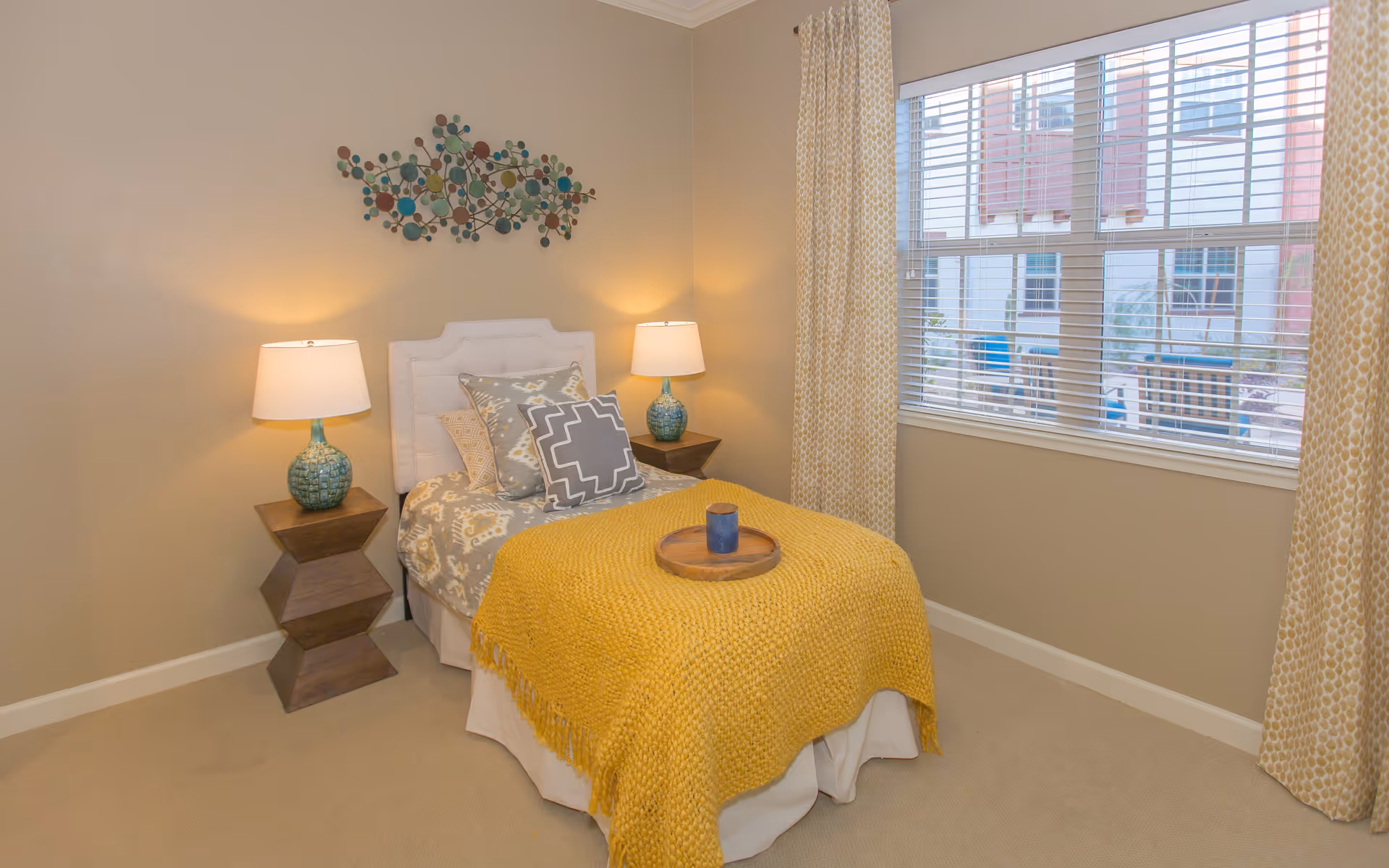 A cozy bedroom with a single bed covered in patterned bedding and a yellow knitted throw blanket. There are three decorative pillows on the bed and a wooden tray with a blue candle on top. Two bedside tables with matching blue lamps flank the bed. A large window with white blinds and yellow patterned curtains lets in natural light. A colorful abstract metal wall art hangs above the bed.