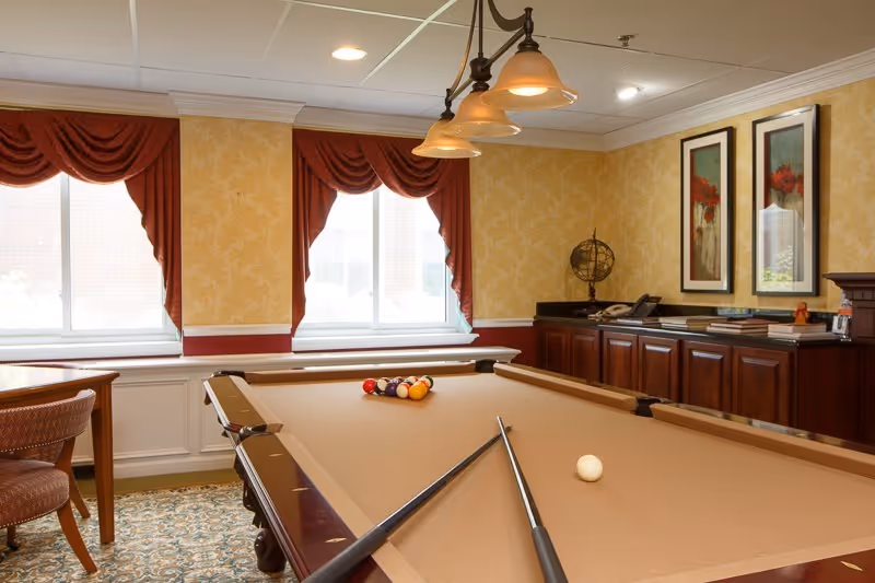 Pool table with cues and racked balls in a warmly decorated game room with draped windows and wood cabinetry.