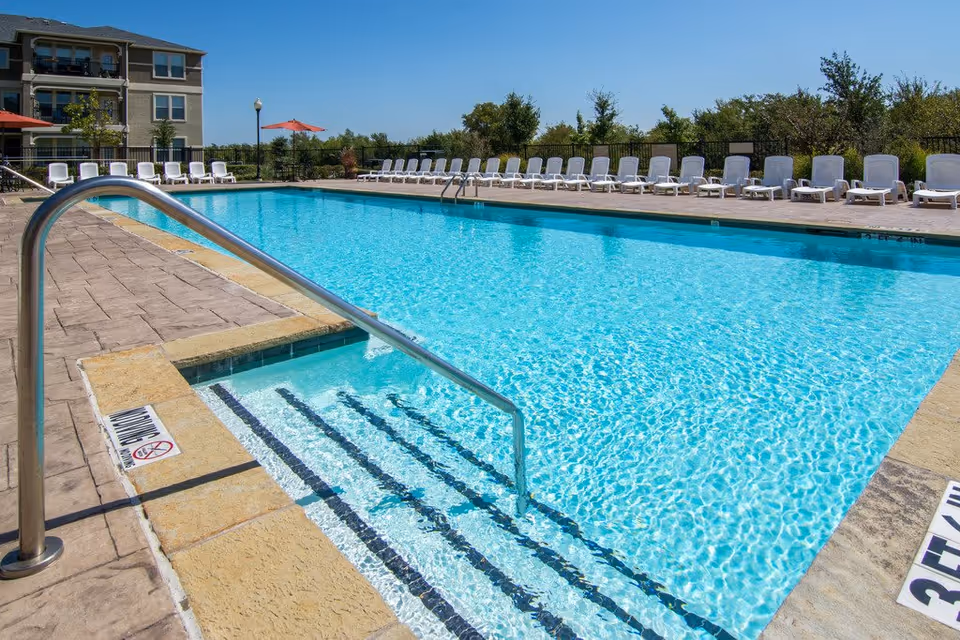 Outdoor swimming pool with entry steps and a metal handrail, surrounded by lounge chairs and a nearby residential building.