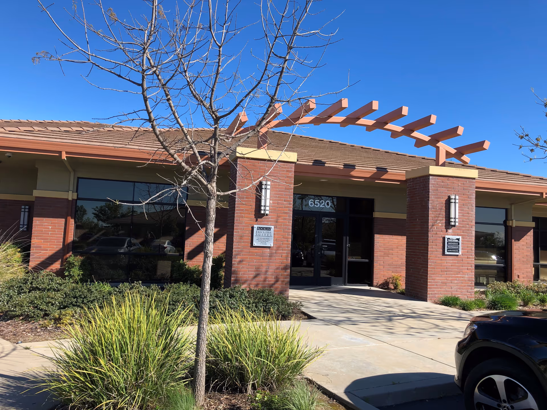 Exterior view of a single-story brick building with large windows and a covered entrance featuring a decorative wooden pergola. There is a small tree and green shrubbery in front of the building under a clear blue sky. A black car is partially visible in the parking area to the right.