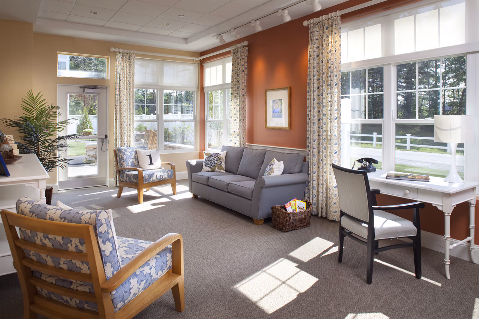 Sunlit communal living room with a gray sofa, patterned armchairs, large windows, and a white desk.