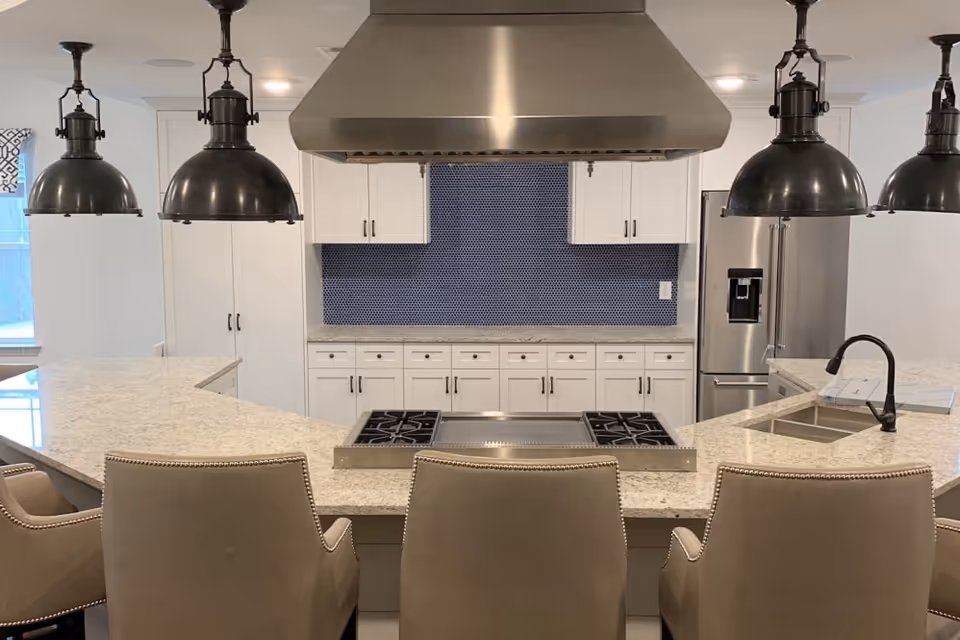 Modern kitchen featuring a large curved granite island with three beige bar stools, industrial pendant lights, a stainless steel range hood and appliances, white cabinetry and a blue tile backsplash.