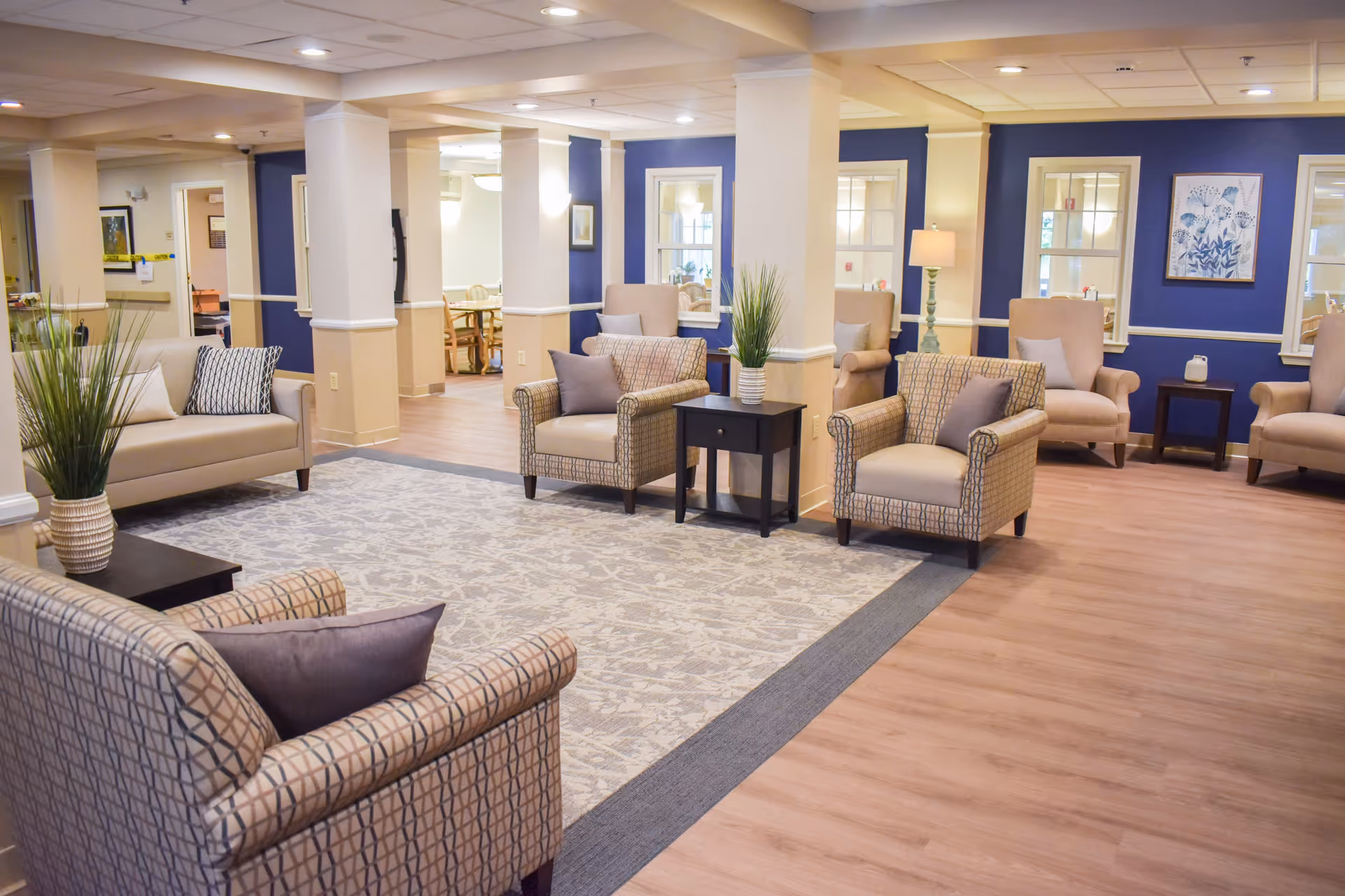 A spacious and well-lit senior living common area with multiple armchairs and sofas arranged around a large patterned rug. The walls are painted blue with white trim, and there are several windows and framed artwork. A small black side table with a potted plant is placed between two armchairs. In the background, a dining area with wooden tables and chairs is visible.