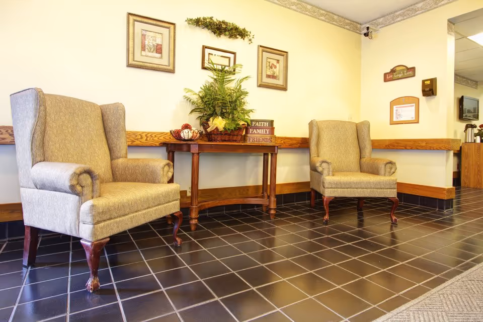 Two upholstered armchairs flank a wooden table topped with a plant and decor beneath framed wall art in a facility seating area.