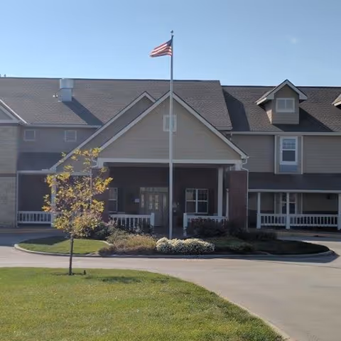 Front exterior view of a senior living facility building with a covered entrance, an American flag on a flagpole, a small tree, and landscaped bushes in front. The building has multiple windows and a gray roof under a clear blue sky.
