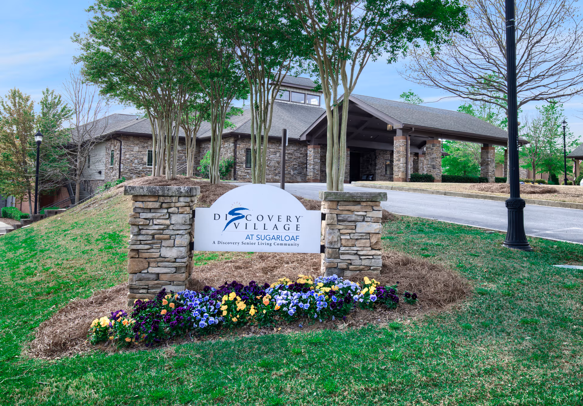 Entrance to Discovery Village at Sugarloaf senior living community featuring a stone sign with flowers in front, a driveway, and a building with a covered entrance surrounded by trees and greenery.