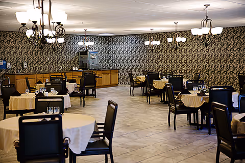 A spacious dining room with multiple round tables covered with beige tablecloths and black chairs arranged around them. The room features patterned wallpaper, tiled flooring, and several chandeliers hanging from the ceiling providing warm lighting. A counter area is visible at the back of the room.
