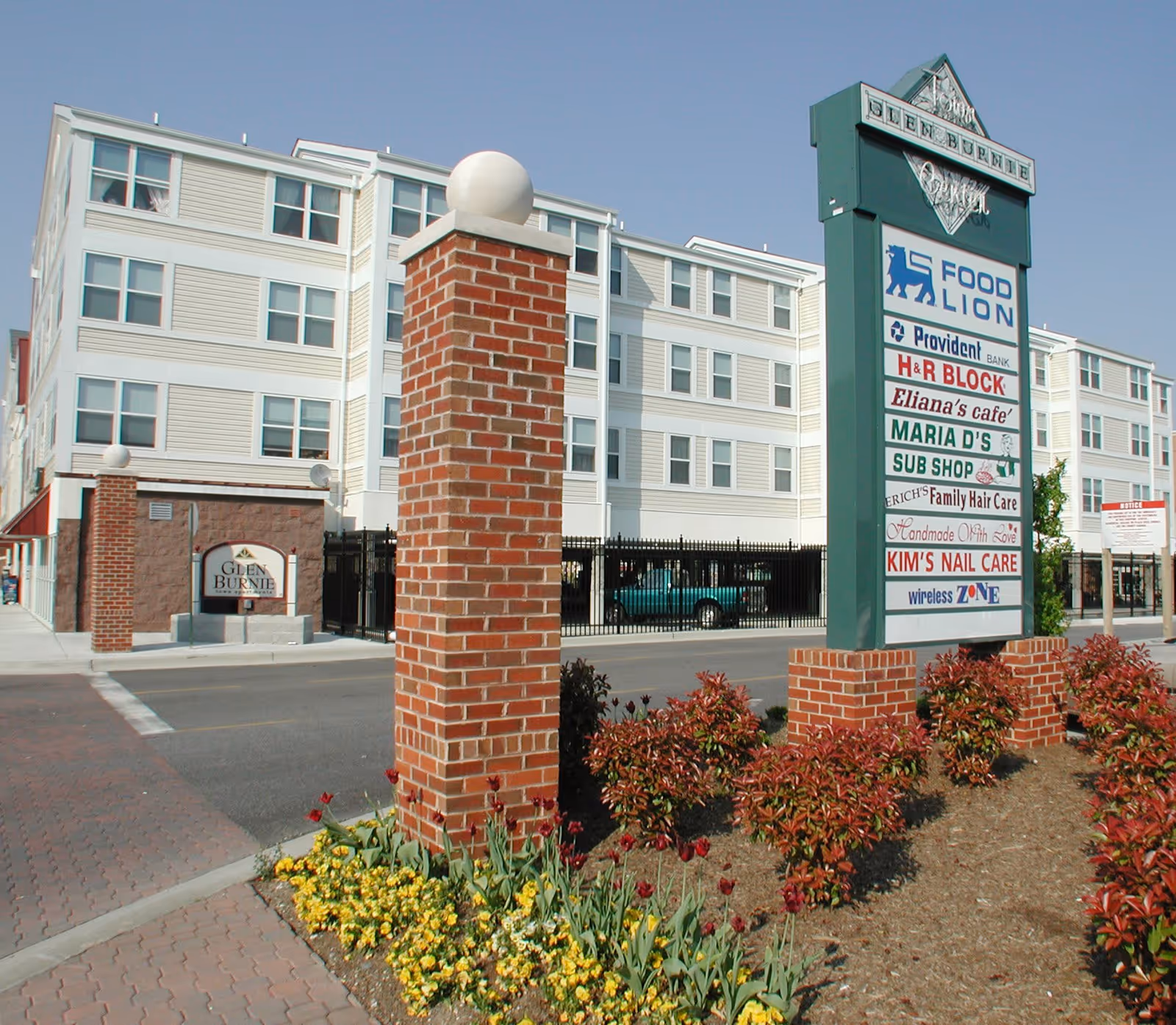 Exterior view of Glen Burnie Town Apartments showing a multi-story residential building with white siding and multiple windows. In the foreground, there is a brick pillar with a white spherical light on top and a landscaped area with flowers and shrubs. A large green sign lists various businesses including Food Lion, Provident Bank, H&R Block, Eliana's Cafe, Maria D's Sub Shop, Erich's Family Hair Care, Kim's Nail Care, and Wireless Zone.