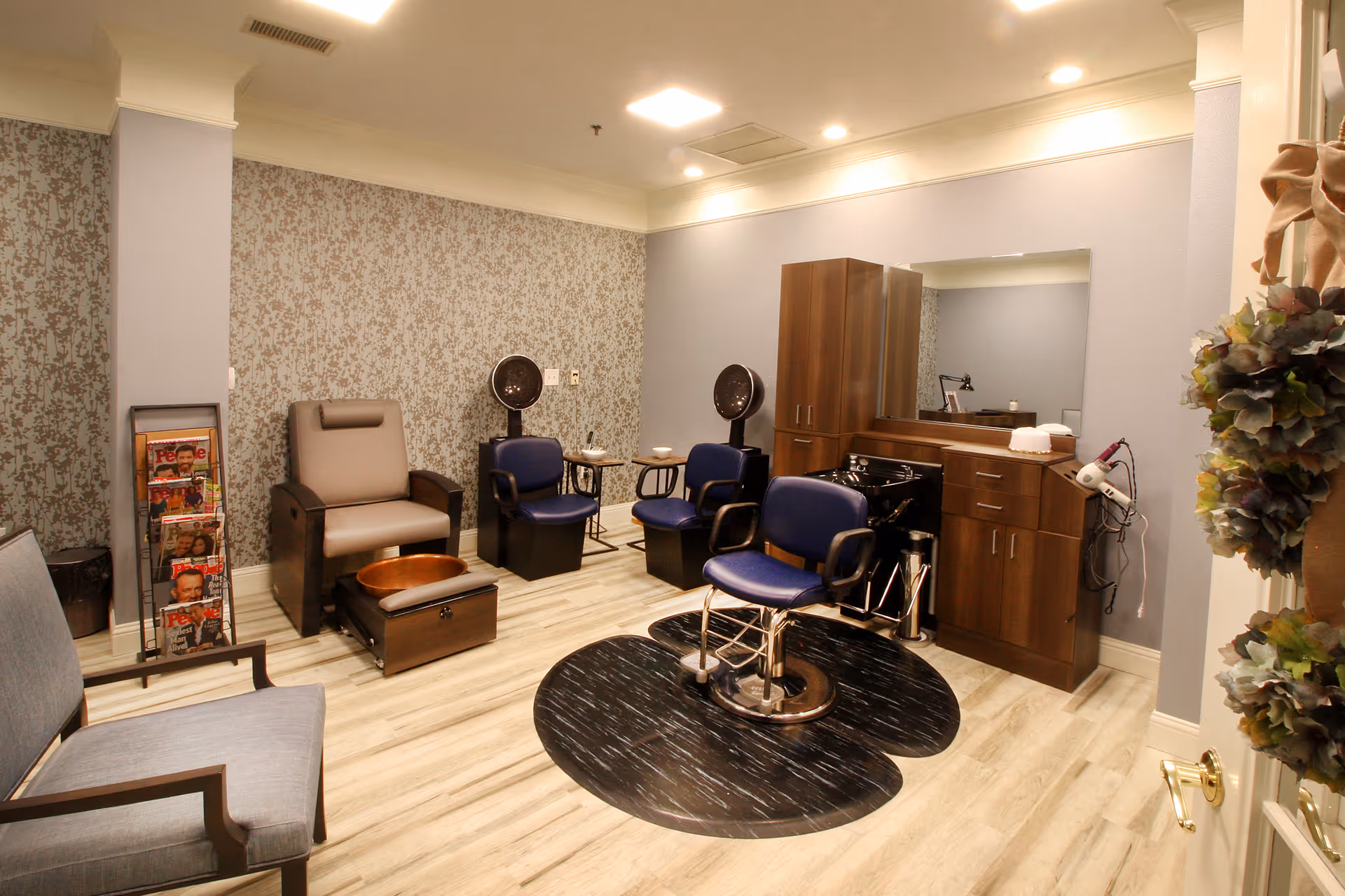 Interior of a senior living facility's salon area with a hair styling chair in front of a large mirror and wooden cabinetry. There are two blue chairs with hair dryers behind them, a gray pedicure chair with a foot bath, a magazine rack with magazines, and additional seating with a blue cushioned chair. The room has light wood flooring and soft lighting.