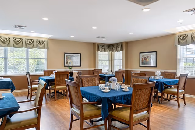 Dining room with several tables covered in blue tablecloths, wooden chairs, place settings, and large windows with valances.
