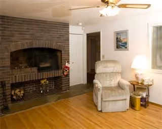 A cozy living room with a brick fireplace on the left, a beige recliner chair, a small wooden side table with a lamp, and a framed picture on the wall. The floor is wooden, and there is a ceiling fan with lights above.