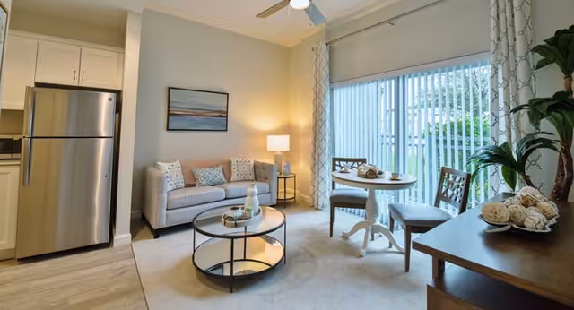 A bright and cozy living area featuring a small gray sofa with decorative pillows, a round glass coffee table, a white round dining table with two chairs, a floor lamp, and a large sliding glass door with vertical blinds leading to an outdoor space. The kitchen area with a stainless steel refrigerator is partially visible on the left, and a wooden sideboard with decorative items is on the right.