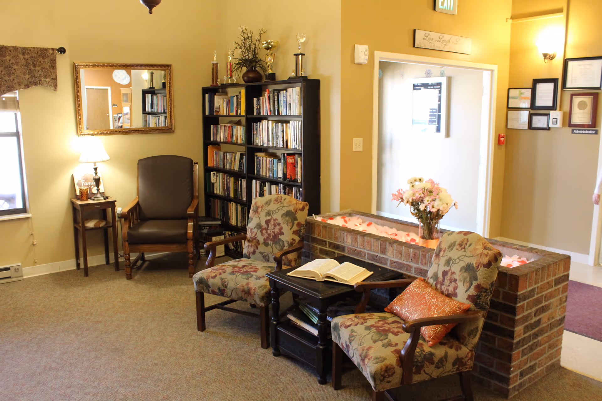 A cozy sitting area in a senior living facility with two floral upholstered armchairs and a small black table holding an open book. Behind the chairs is a low brick planter with flowers. To the left, there is a brown armchair next to a small side table with a lamp and decorative items. A tall black bookshelf filled with books and trophies stands against the wall. The walls are painted beige, and there is a mirror hanging above the brown armchair. An exit door is visible in the background with certificates and a sign labeled 'Administrator' on the wall nearby.