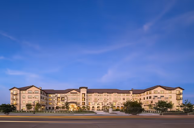 Wide exterior view of a large, multi-story senior living facility building under a clear blue sky at dusk, with well-lit windows and landscaped greenery in front.