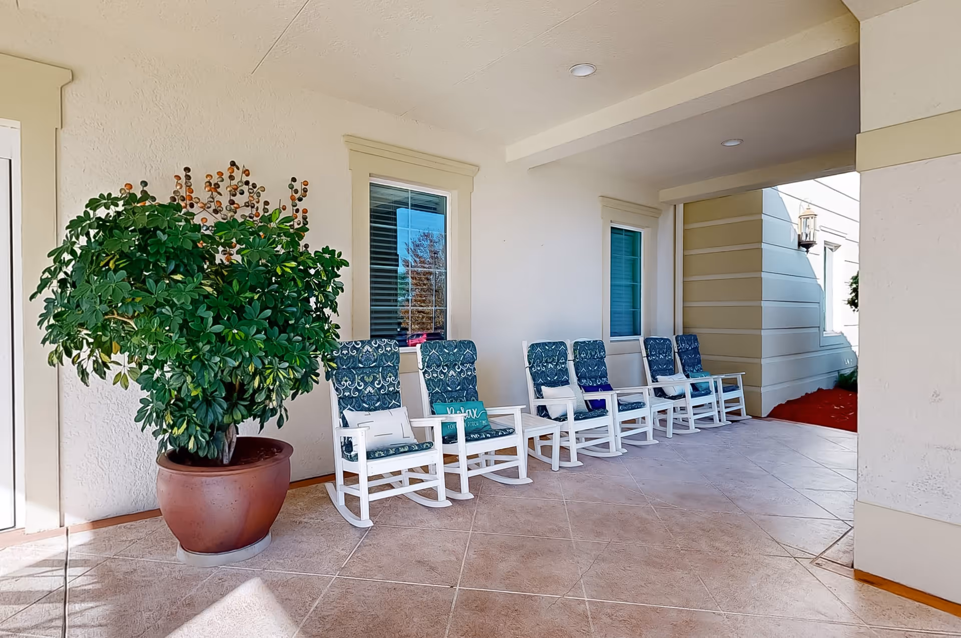 A covered outdoor patio area with six white rocking chairs lined up against a beige wall with two windows. Each chair has a blue patterned cushion and some have small pillows. A large potted plant with green leaves is placed on the left side near the chairs. The floor is tiled and the area is well-lit with natural light.