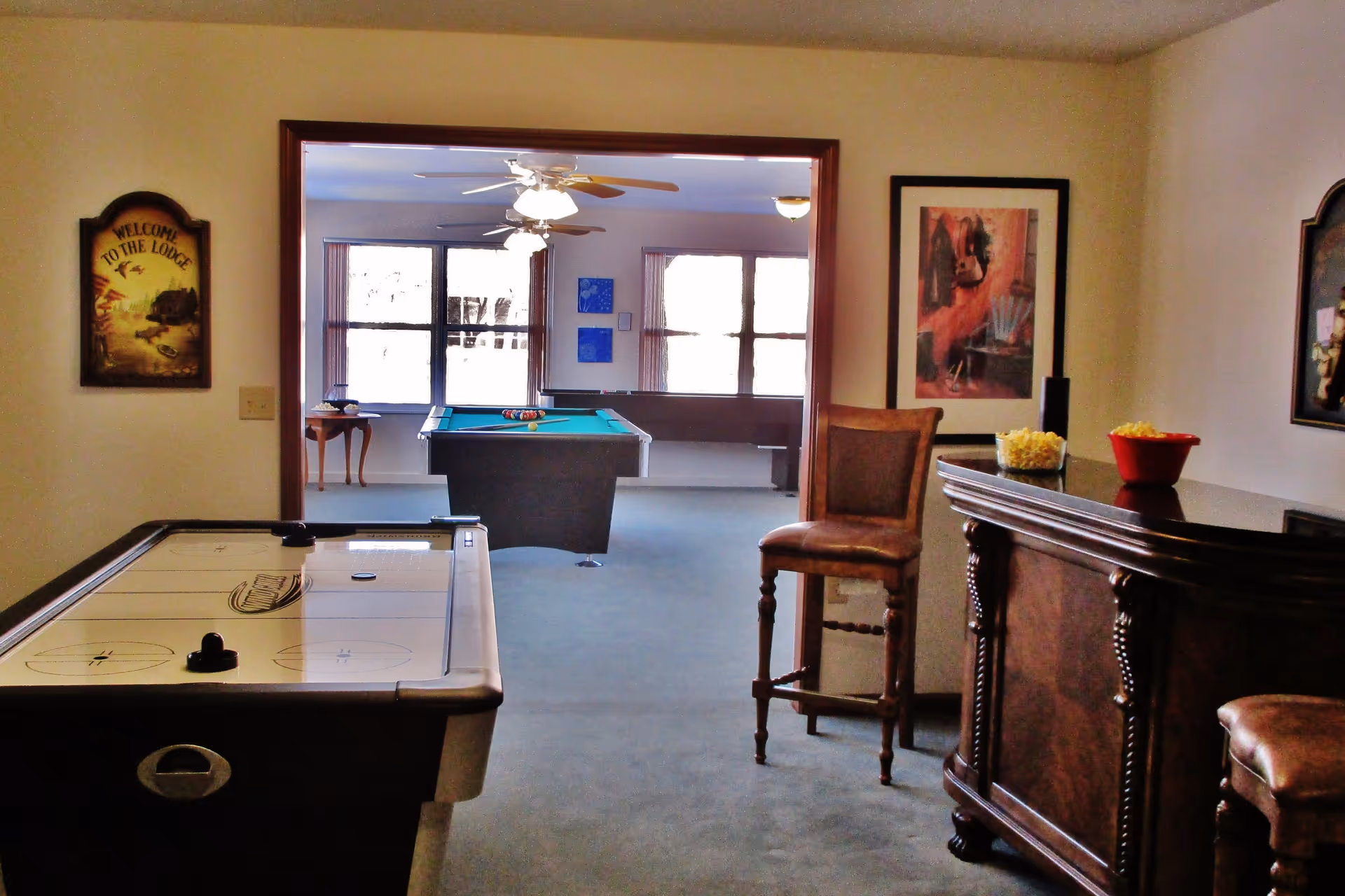 Interior view of a recreational lounge area featuring an air hockey table in the foreground, a wooden bar with stools on the right, and a pool table in the adjacent room with large windows letting in natural light. Wall decorations include framed artwork and a sign that reads 'Welcome to the Lodge'.
