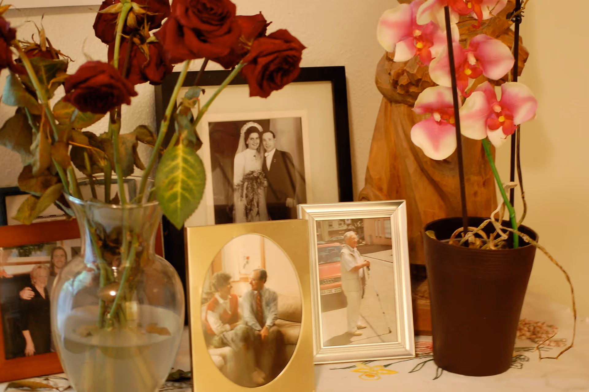 Tabletop display with framed family photographs, a vase of roses and a potted orchid on a cloth-covered surface.