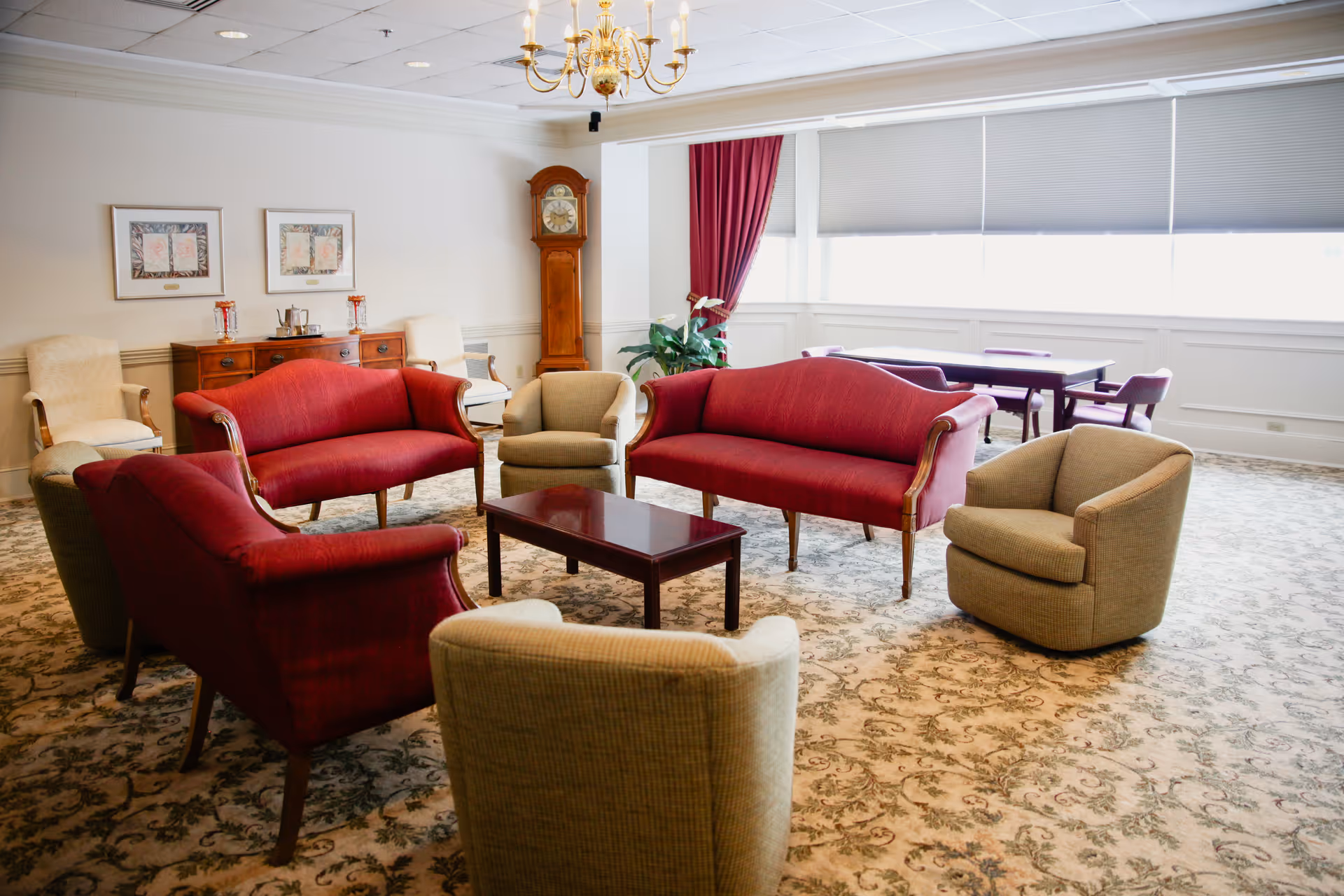 A spacious living room with red upholstered sofas and beige armchairs arranged around a wooden coffee table. The room features a patterned carpet, a wooden grandfather clock, a sideboard with decorative items, framed artwork on the wall, a chandelier, and large windows with blinds and red curtains.