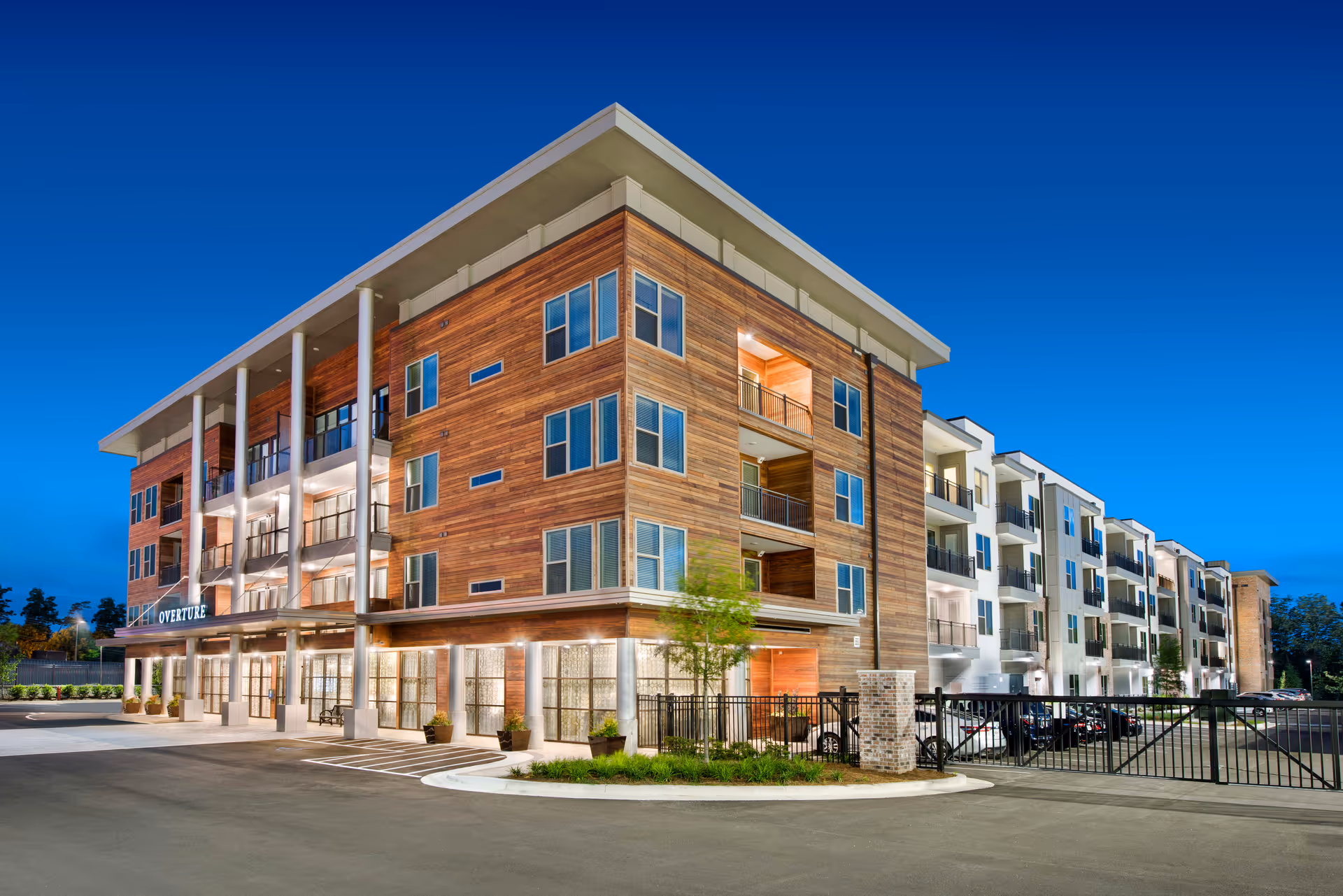 Exterior view of a modern multi-story senior living facility named Overture Barrett at dusk, featuring wood paneling and balconies, with a gated parking area in front.