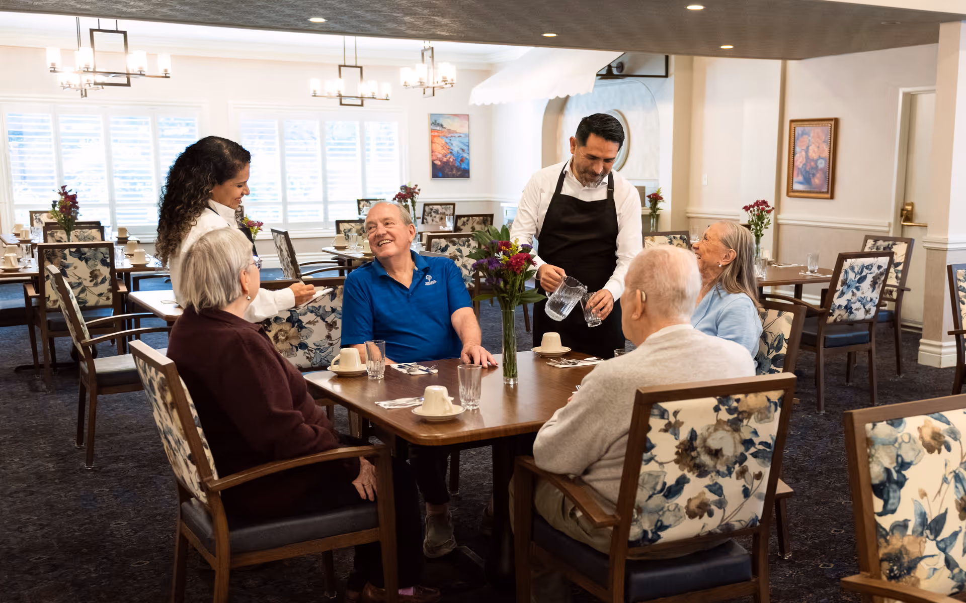 A group of elderly people sitting around a dining table in a well-lit dining room with floral upholstered chairs. A female staff member is taking an order from one of the residents, while a male staff member is pouring water into a glass. The room has large windows with white shutters, framed artwork on the walls, and flower arrangements on the tables.