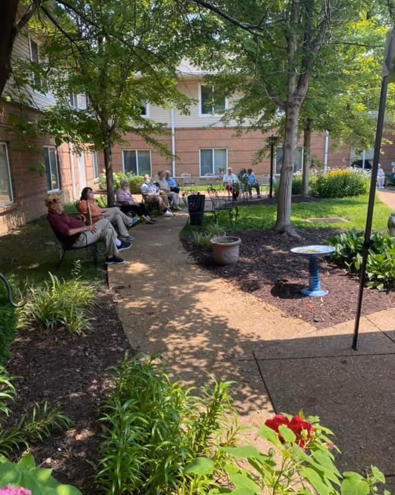 A group of elderly people sitting on benches along a curved pathway in a shaded garden courtyard with trees and plants, adjacent to a brick building with windows.