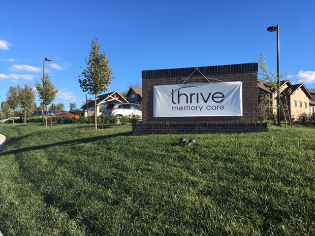 Outdoor view of a grassy area with a brick sign displaying 'thrive memory care' in front of a building complex under a blue sky with scattered clouds.