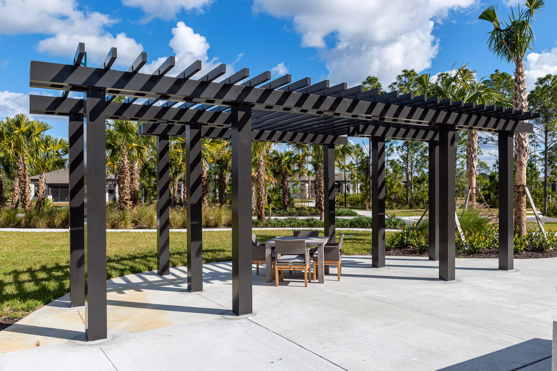Outdoor seating area under a modern black pergola with a table and four chairs on a concrete patio surrounded by grass, palm trees, and other greenery under a partly cloudy blue sky.