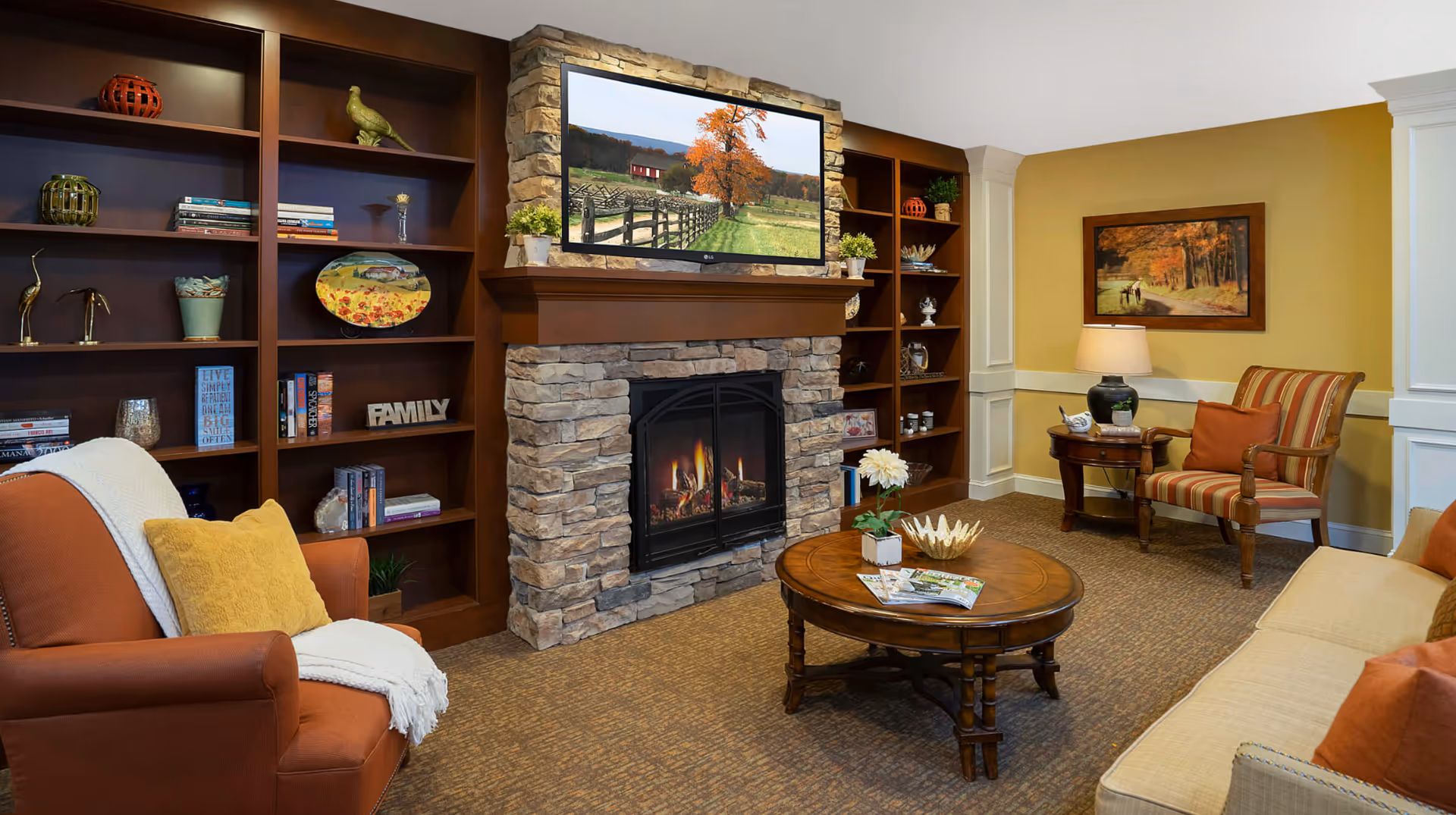 A cozy living room with a stone fireplace and a flat-screen TV mounted above it displaying a scenic autumn landscape. The room features built-in wooden shelves filled with books and decorative items, a round wooden coffee table with magazines and a flower vase, an orange armchair with a yellow pillow and white throw, a striped armchair next to a side table with a lamp, and a beige sofa with orange pillows. The walls are painted yellow with white trim and a framed autumn-themed painting hangs on the wall.
