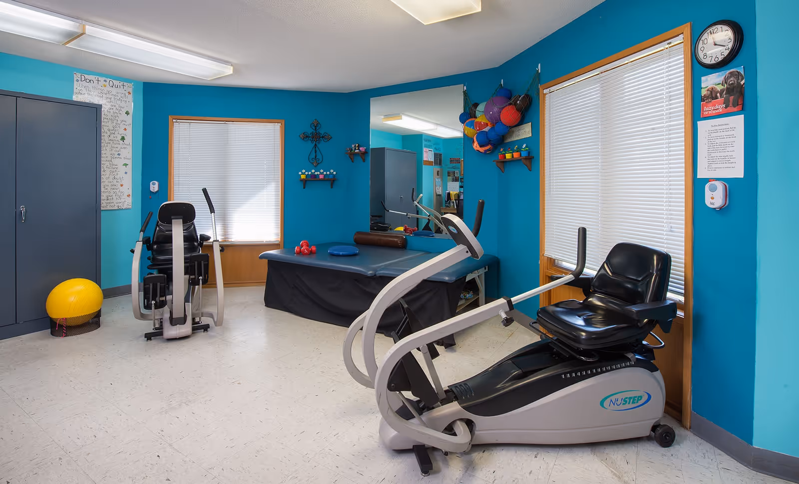 Bright teal-walled therapy/exercise room with two recumbent machines, a treatment table, fitness balls, and storage cabinets.