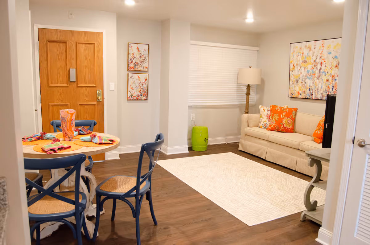 A cozy senior living community interior featuring a small round dining table set with colorful napkins and placemats, surrounded by four blue chairs. Adjacent to the dining area is a living room with a beige sofa adorned with orange and floral cushions, a floor lamp, a green decorative stool, and a large abstract painting on the wall. The room has wooden flooring and a light-colored area rug.