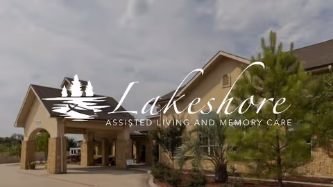 Exterior view of Lakeshore Assisted Living and Memory Care building featuring a covered entrance with stone pillars, surrounded by trees and landscaping under a partly cloudy sky.