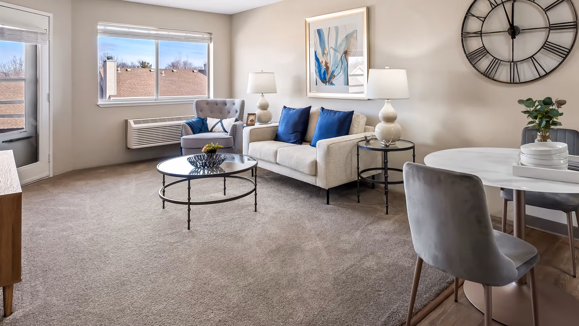 A bright living room with beige carpet and neutral walls featuring a beige sofa with blue pillows, a gray armchair, a round glass coffee table, two side tables with white lamps, a round dining table with gray chairs, a large wall clock, and a window showing rooftops outside.