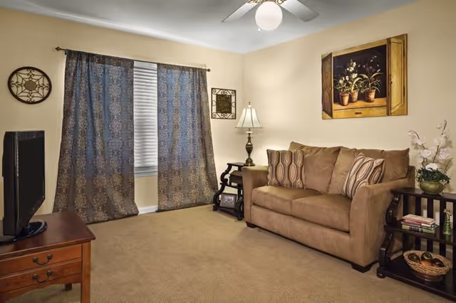 Cozy living room with a brown sofa, patterned curtains over a window, side tables with a lamp and a TV on a wooden stand.
