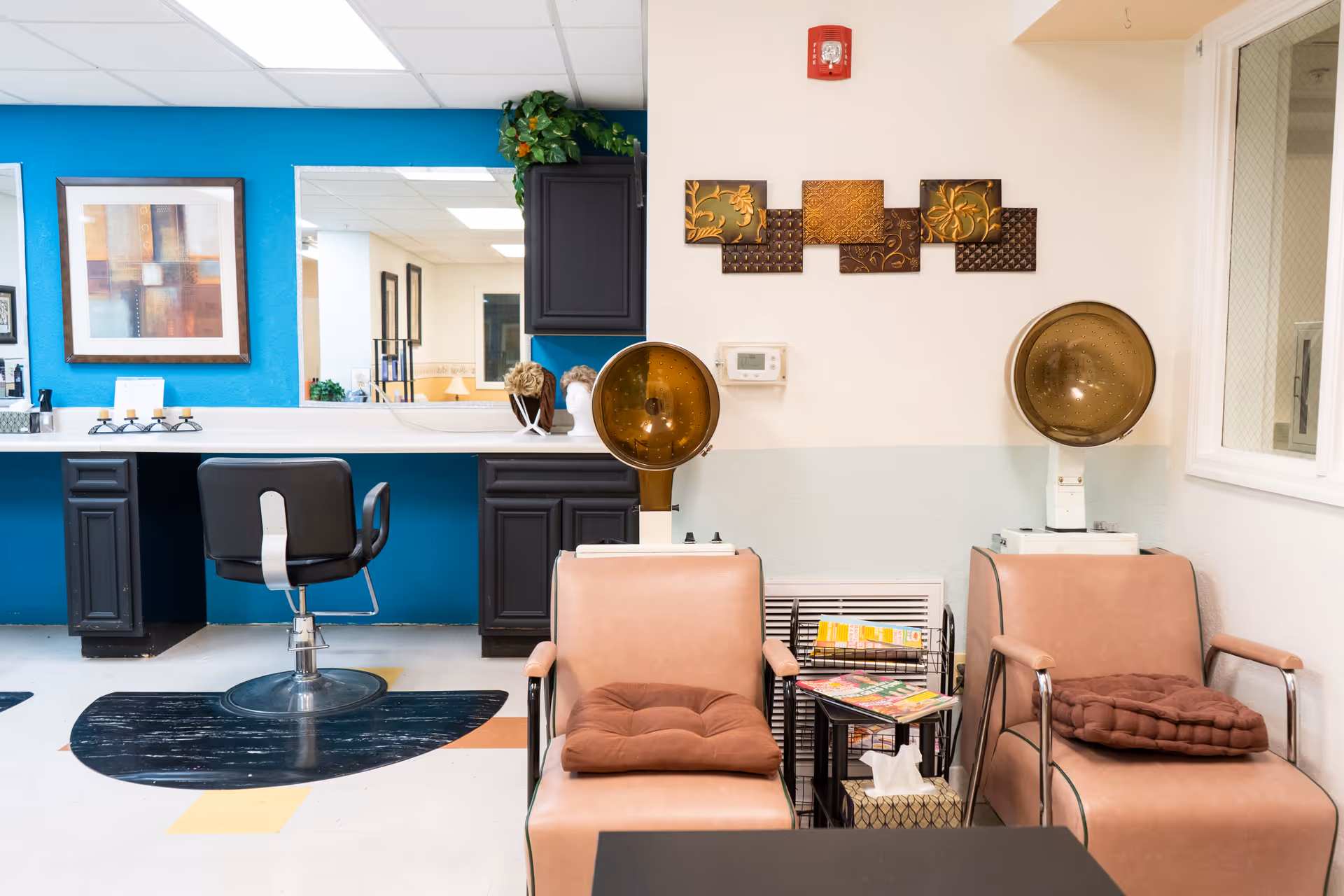 Interior view of a salon area in a senior living facility with two vintage hair dryers and two cushioned chairs. There is a black salon chair in front of a counter with a large mirror and blue accent wall. Decorative artwork and plants are visible on the walls and counter.