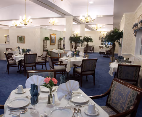Formal dining room with tables set with white linens, folded napkins, china, chandeliers, and decorative plants.