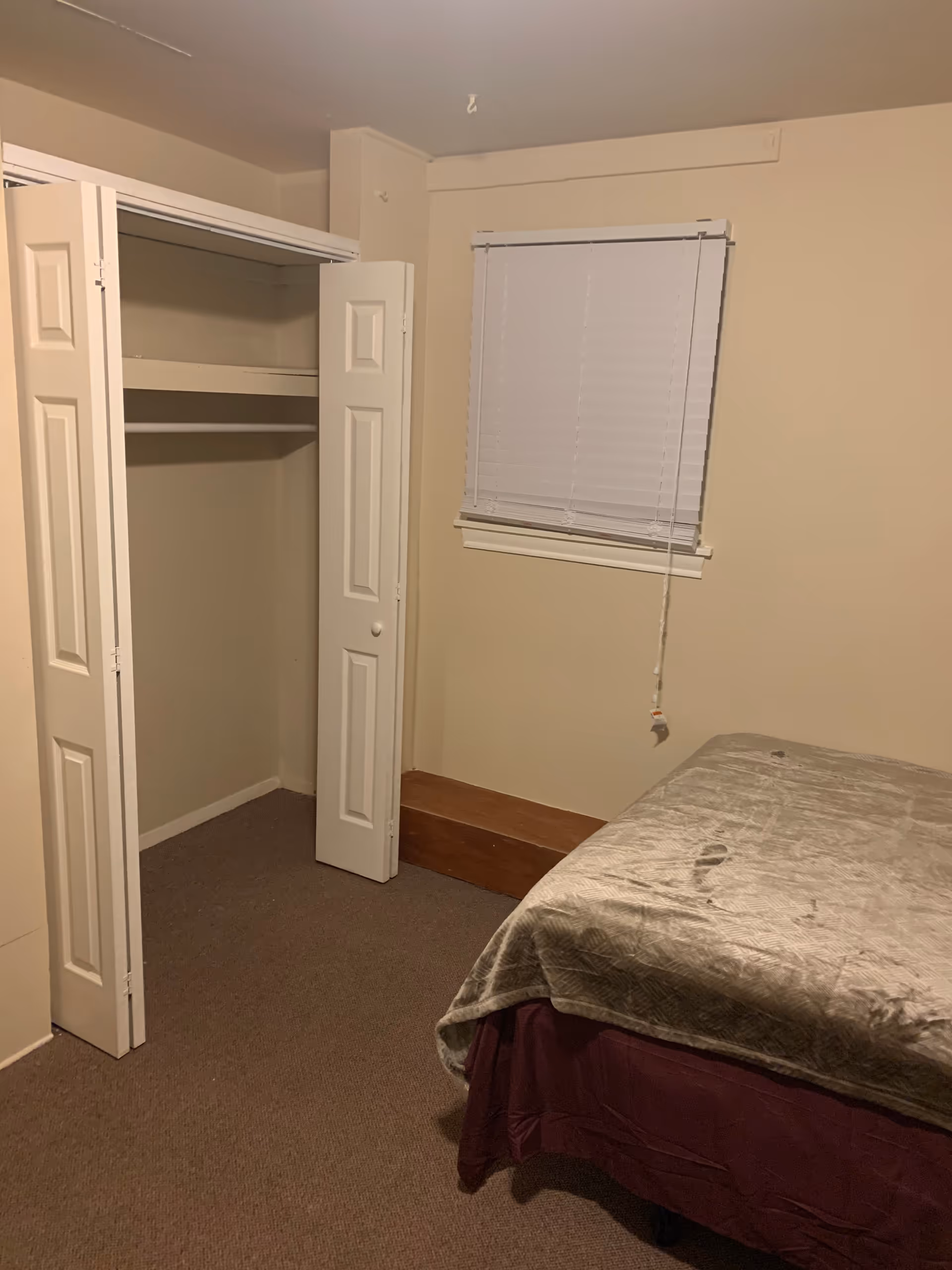 A small bedroom with beige walls and carpeted floor. There is a bed with a gray blanket and a maroon bed skirt on the right side. On the left side, there is an open closet with white bi-fold doors. A window with closed white blinds is centered on the wall above a wooden ledge.