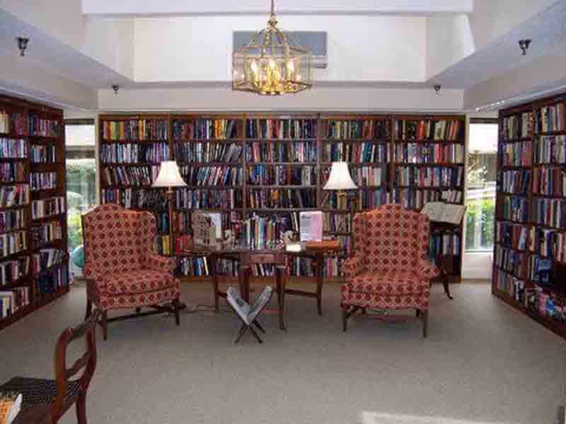 A cozy library room with tall wooden bookshelves filled with books lining the walls. Two patterned armchairs face each other with a small wooden table between them, holding lamps and books. A chandelier hangs from the ceiling, and windows on either side let in natural light.