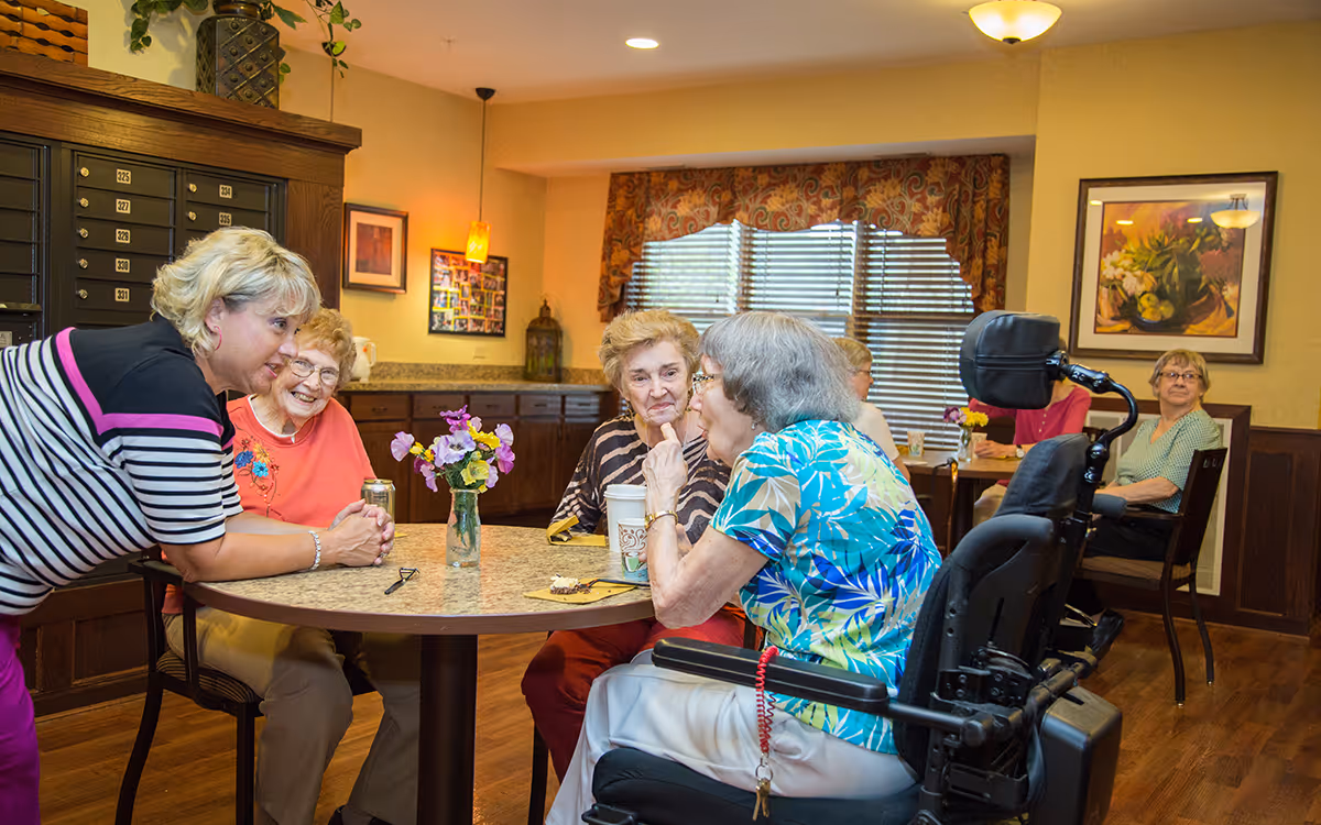 A group of elderly women sitting and chatting around a round table in a warmly lit common area. One woman in a wheelchair is engaged in conversation with others. The room has wooden flooring, decorative curtains, framed artwork on the walls, and a vase with flowers on the table.