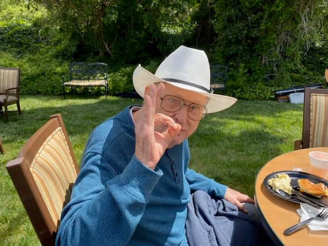 An elderly man wearing a white hat and glasses sits outdoors at a round table with a plate of food. He is making an OK hand gesture and smiling. The background shows green grass, trees, and outdoor chairs.