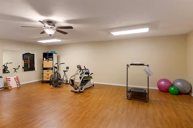 A small fitness room with exercise bikes, a treadmill, stability balls, dumbbells and a wall mirror on wood flooring.