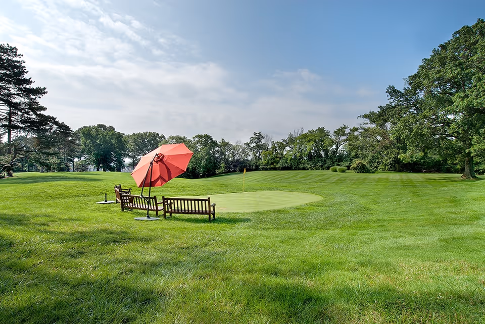 A well-maintained green lawn with a small putting green and a yellow flag. Two wooden benches and a large red umbrella are placed near the putting green. Trees and shrubs surround the area under a partly cloudy blue sky.
